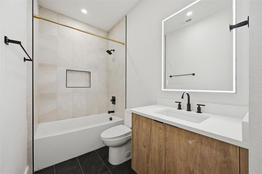 Secondary Bathroom features recessed lighting. Large-format tile surrounds the soaking tub and shower area. Wood vanity includes quartz countertop beneath a backlit mirror. Black fixtures accent the space. Tile flooring extends throughout.
