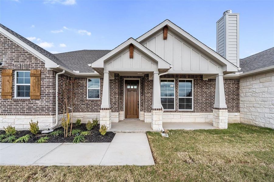 View of exterior entry featuring a yard, brick siding, a shingled roof, a porch, and board and batten siding View of exterior entry featuring a yard, brick siding, a shingled roof, a porch, and board and batten siding