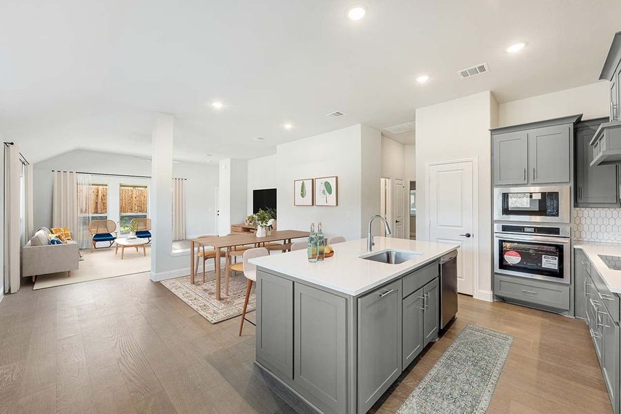 Open-concept kitchen with gray cabinets, center island with sink, and view into dining and living areas with hardwood floors