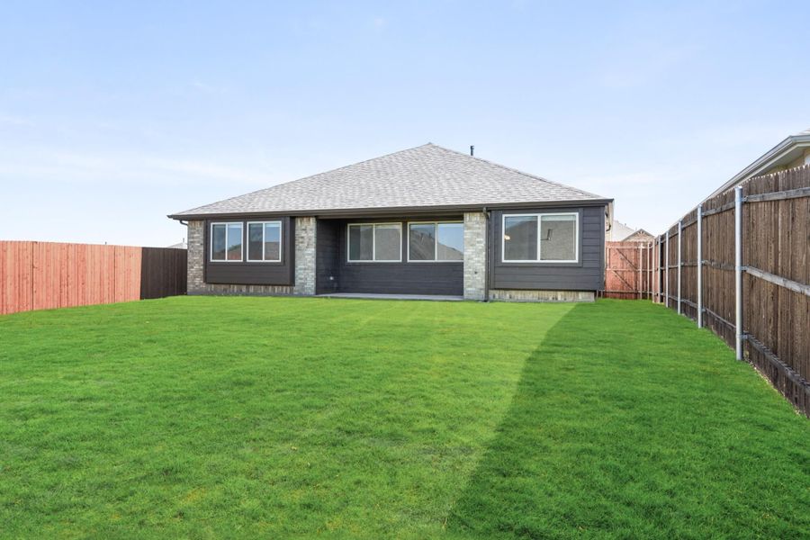 Rear view of single-story home with dark siding, brick accents, and large fenced backyard with green lawn