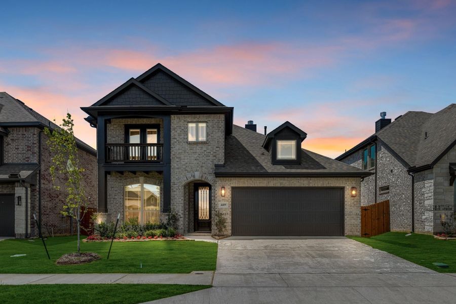 Two-story brick home with dark trim, second-floor balcony, two-car garage, and landscaped front yard at dusk
