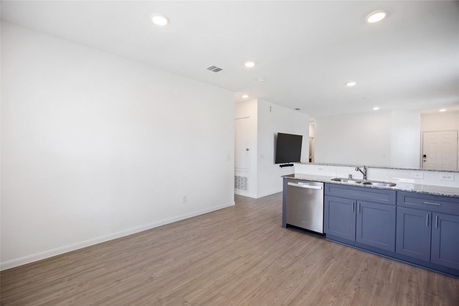 bright, open-concept living space with light wood flooring and recessed lighting. The modern kitchen features a dark blue island with a double sink and stainless steel dishwasher, seamlessly integrating with the living area.