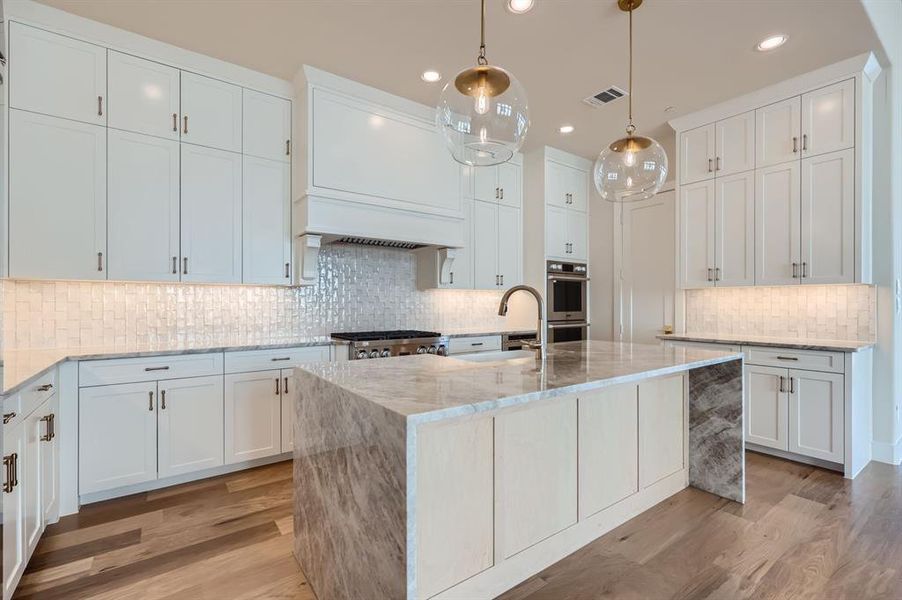 Kitchen featuring light wood-type flooring, pendant lighting, white cabinetry, and a kitchen island with sink