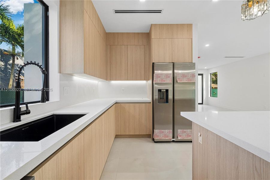 Kitchen featuring flat-panel wood-finish cabinetry, white countertops, black undermount sink with high-arc faucet, stainless steel refrigerator, and recessed lighting Kitchen featuring flat-panel wood-finish cabinetry, white countertops, black undermount sink with high-arc faucet, stainless steel refrigerator, and recessed lighting