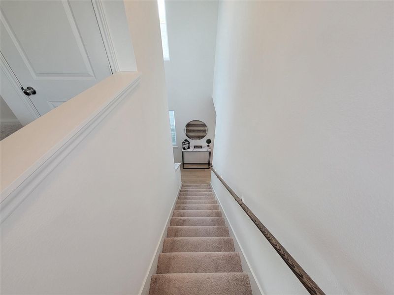 A well-lit staircase with carpeted steps and a wooden handrail leads down to a landing area. The walls are painted white, giving a clean and modern look.