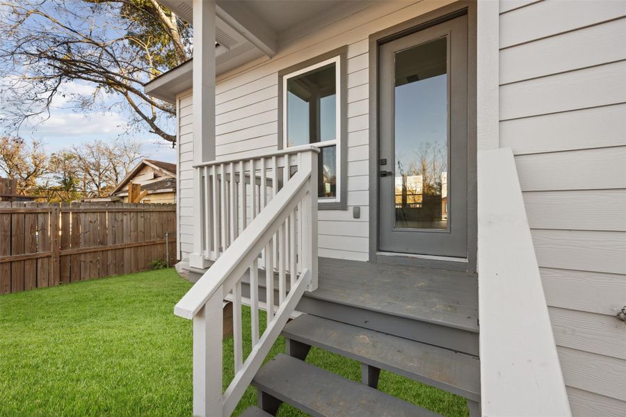 This photo shows a small back porch with steps leading to a fenced yard. The exterior features light-colored siding, a glass-paneled door, and a well-maintained lawn, providing a cozy outdoor space. This photo shows a small back porch with steps leading to a fenced yard. The exterior features light-colored siding, a glass-paneled door, and a well-maintained lawn, providing a cozy outdoor space.