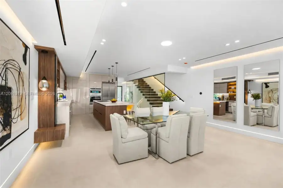 Modern kitchen and dining area featuring high-gloss cabinetry, wood-finish wall paneling with integrated lighting, a stone-top kitchen island, recessed linear ceiling lighting, and a floating wood-tread staircase with a glass railing Modern kitchen and dining area featuring high-gloss cabinetry, wood-finish wall paneling with integrated lighting, a stone-top kitchen island, recessed linear ceiling lighting, and a floating wood-tread staircase with a glass railing