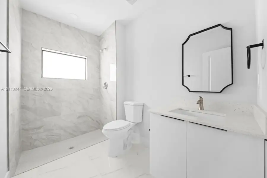 Full bathroom featuring a walk-in shower with stone-finish tile surround, white vanity cabinet with quartz countertop, undermount sink, and geometric wall-mounted mirror Full bathroom featuring a walk-in shower with stone-finish tile surround, white vanity cabinet with quartz countertop, undermount sink, and geometric wall-mounted mirror
