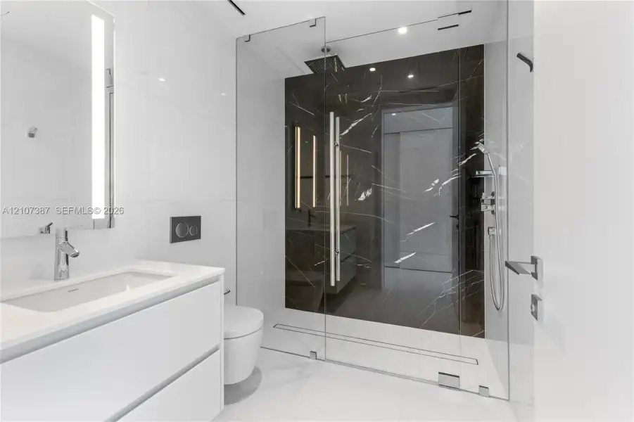 Modern bathroom featuring a floating white vanity with a rectangular sink, wall-mounted faucet, walk-in shower with floor-to-ceiling glass enclosure, dark stone-finish shower surround, and integrated lighting
