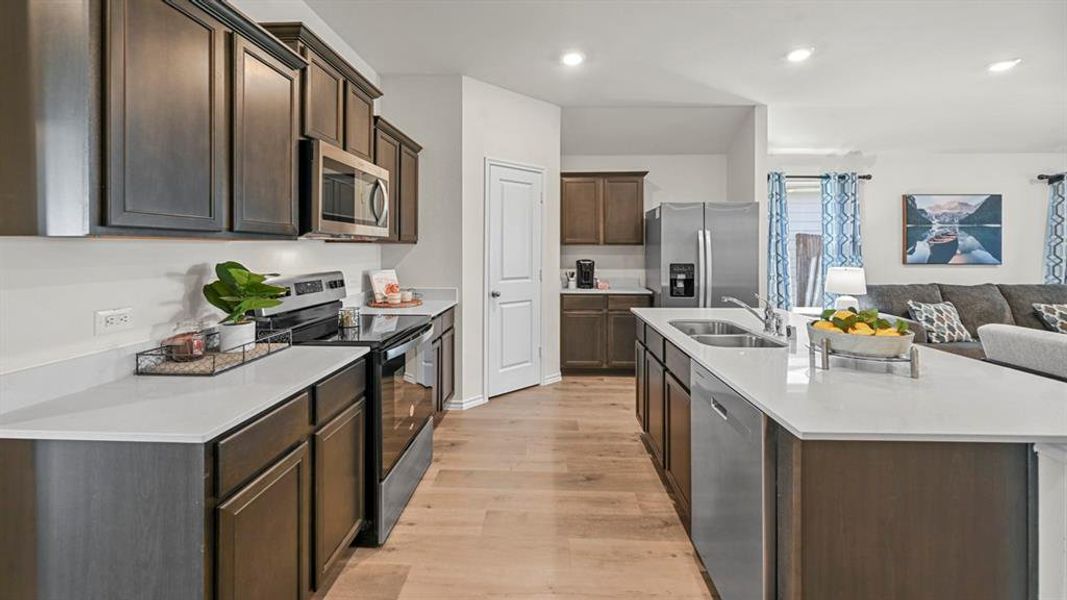 Kitchen with stainless steel appliances, dark wood finish cabinetry, a center island with sink, open floor plan, and light wood-style floors