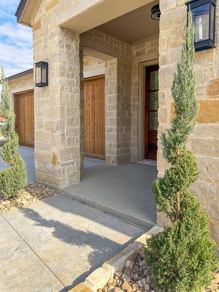 Stone facade entry with covered porch featuring wood grain garage doors, a dark wood entry door, and decorative sconce lighting