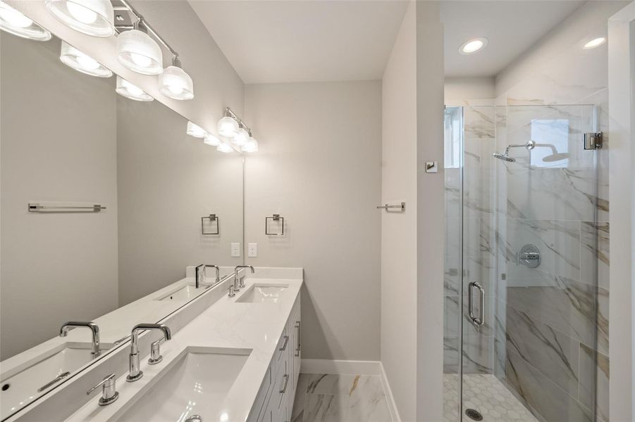 This modern bathroom features a double vanity with sleek faucets, ample lighting, and a spacious glass-enclosed shower with elegant marble-style tiles.