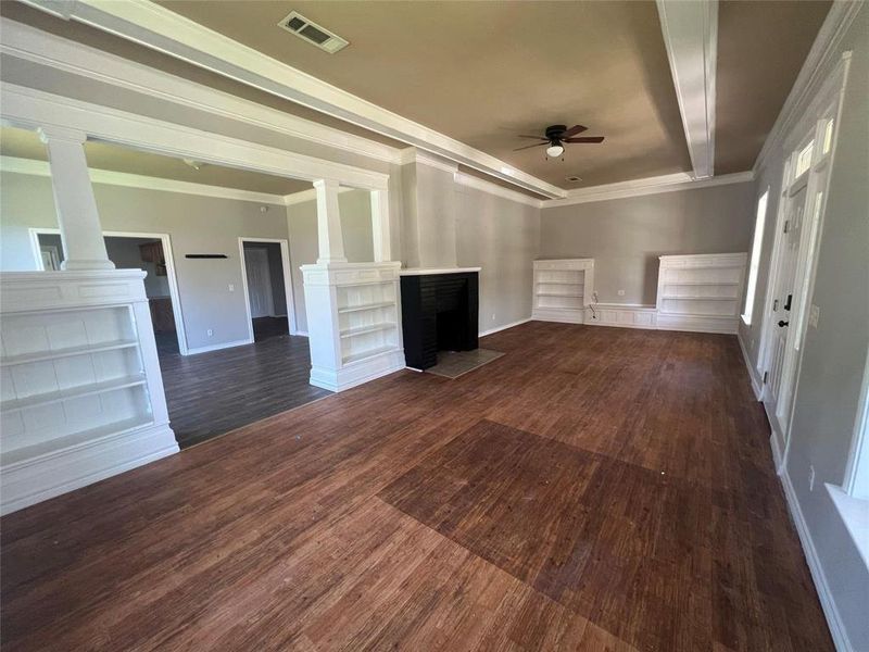 Unfurnished living room with a fireplace, crown molding, a ceiling fan, dark wood-type flooring, and built in shelves Unfurnished living room with a fireplace, crown molding, a ceiling fan, dark wood-type flooring, and built in shelves