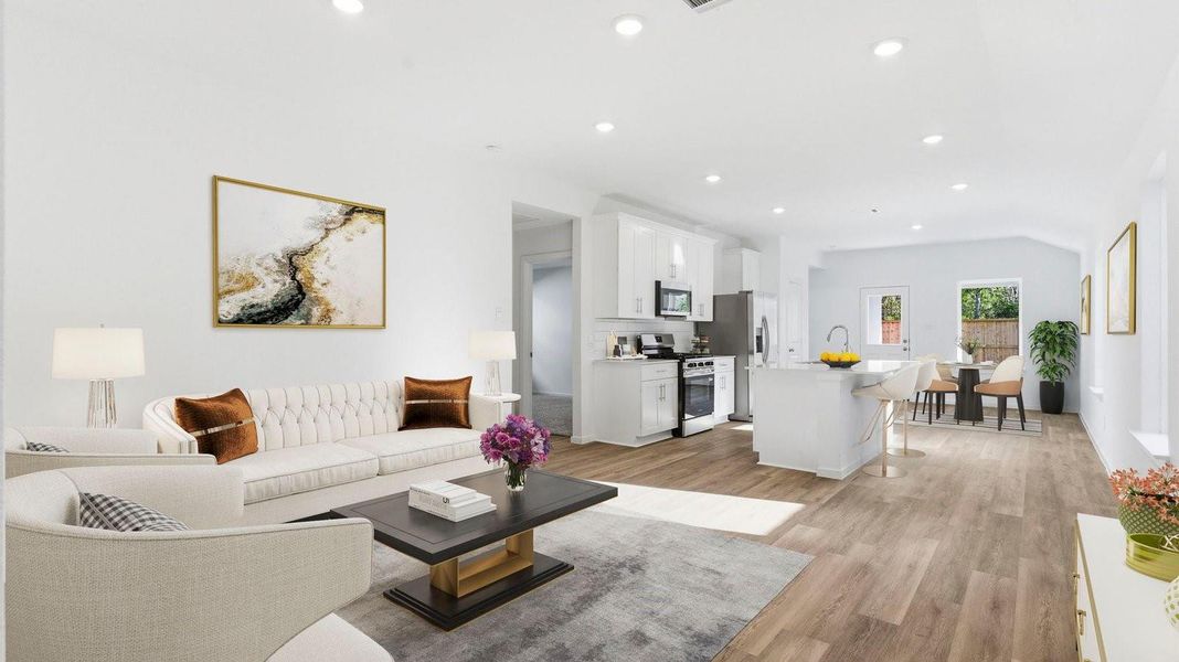 A cozy living room featuring large windows for natural lighting, wood flooring and a view to the kitchen and dining area.