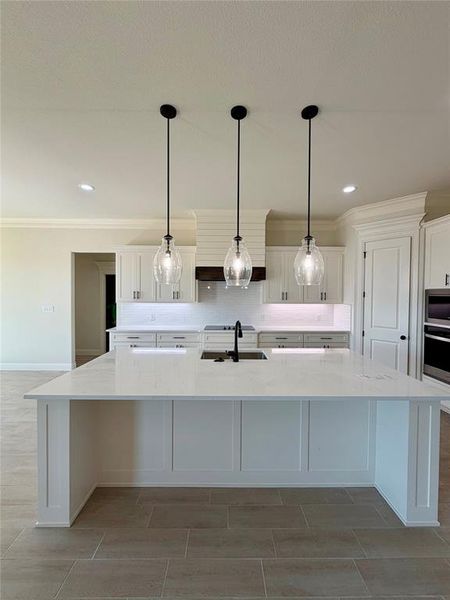 Kitchen with white cabinets, a spacious island, light stone countertops, decorative light fixtures, and ornamental molding Kitchen with white cabinets, a spacious island, light stone countertops, decorative light fixtures, and ornamental molding
