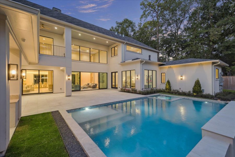 Beautiful modern pool featuring a spacious design with a flushed spa, elegant tile accents, and a sleek water wall. Expansive stone decking surrounds the pool, offering ample space for lounging and entertaining, while the backdrop of large windows and architectural lines creates a seamless indoor–outdoor connection.