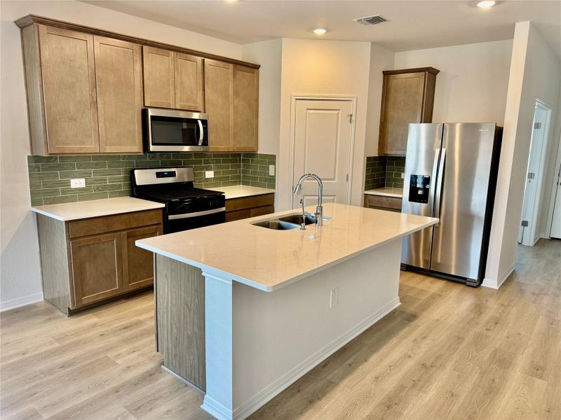 Kitchen featuring appliances with stainless steel finishes, backsplash, a center island with sink, light stone counters, and light wood-type flooring Kitchen featuring appliances with stainless steel finishes, backsplash, a center island with sink, light stone counters, and light wood-type flooring