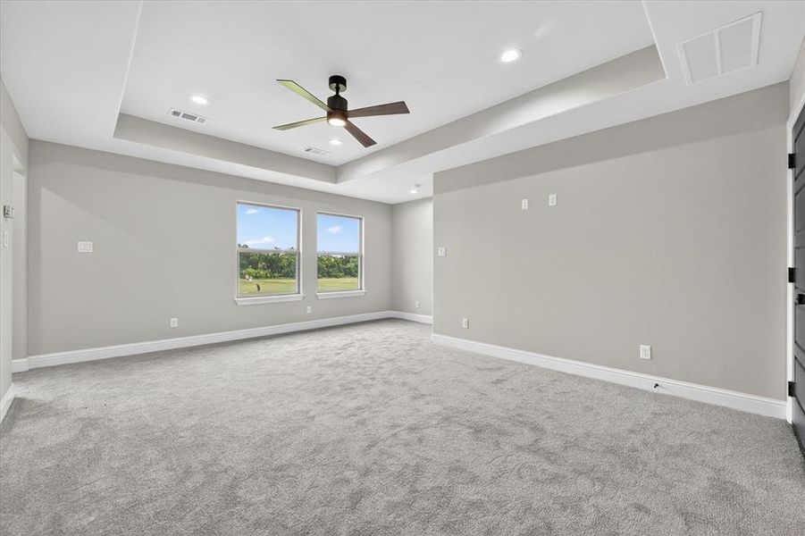 Spacious room featuring light gray carpeting, two large windows, a tray ceiling with recessed lighting, and a dark-toned ceiling fan
