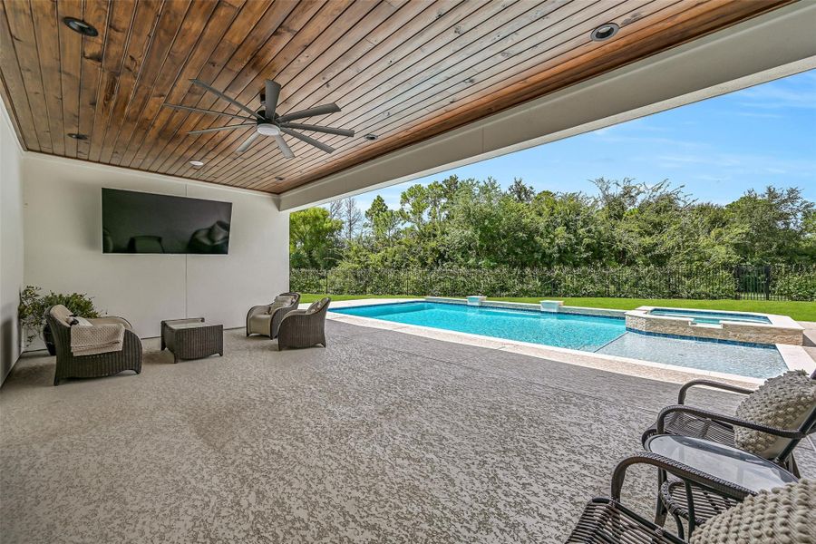 A huge covered patio dramatically extends the living space outdoors with a warm wood-plank ceiling, recessed lighting, oversized fan & mounted TV, all overlooking the Aqua Sport pool, raised spa and private tree-lined backdrop.