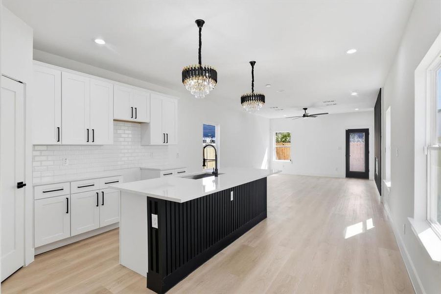 Open-concept kitchen and living area featuring white shaker cabinetry, an island with slatted black millwork, light wood-finish flooring, recessed lighting, and stylish chandeliers