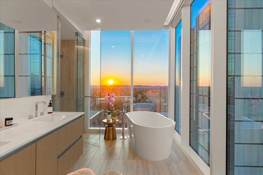 Soak in luxury with breathtaking sunset views from this spa-inspired primary bath. A freestanding soaking tub is framed by floor-to-ceiling windows, while the floating double vanity features custom marble counters and modern cabinetry. A glass-enclosed walk-in shower completes the composition. This serene retreat blends elevated design with panoramic skyline and Hill Country vistas.
