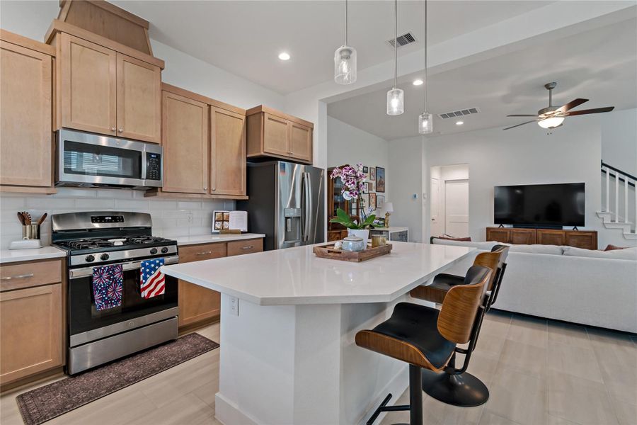 Modern kitchen with light wood cabinetry, stainless steel appliances, and a spacious island with seating, seamlessly flowing into an open-concept living area featuring a cozy sofa and entertainment setup. Modern kitchen with light wood cabinetry, stainless steel appliances, and a spacious island with seating, seamlessly flowing into an open-concept living area featuring a cozy sofa and entertainment setup.