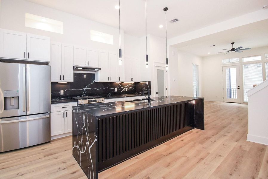 This spacious, modern kitchen features sleek black marble countertops with a matching backsplash, stainless steel appliances, and white cabinetry. The open layout flows into a bright living area with large windows and a ceiling fan, complemented by stylish pendant lighting and light wood flooring.
