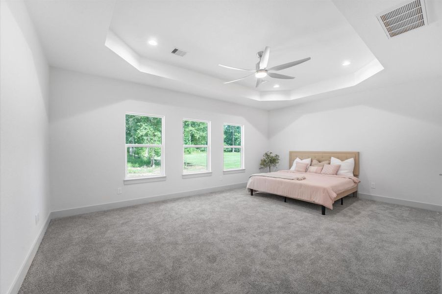 This spacious primary bedroom features a tray ceiling with recessed lighting and a modern ceiling fan. It has three large windows offering plenty of natural light and a serene view of greenery. The room is carpeted and includes a neatly placed bed, providing a cozy and inviting atmosphere. This spacious primary bedroom features a tray ceiling with recessed lighting and a modern ceiling fan. It has three large windows offering plenty of natural light and a serene view of greenery. The room is carpeted and includes a neatly placed bed, providing a cozy and inviting atmosphere.
