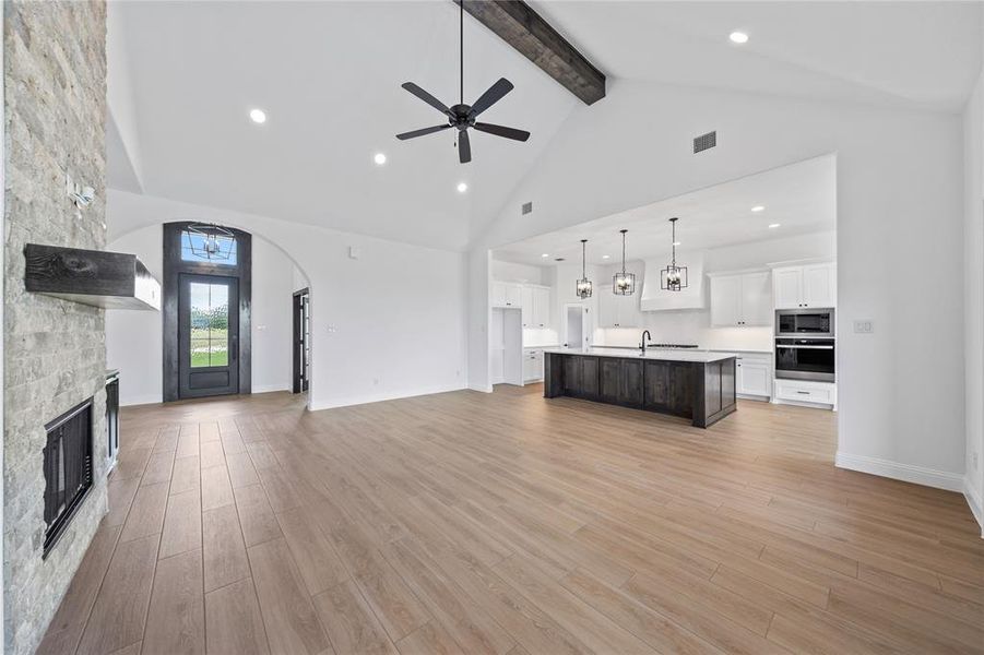 Unfurnished living room featuring high vaulted ceiling, a ceiling fan, beam ceiling, a stone fireplace, and light wood finished floors