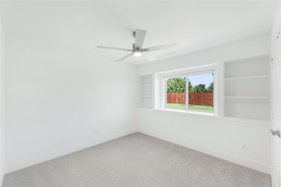 Empty bedroom with carpet, ceiling fan, built-in shelves, and a window overlooking a fenced backyard