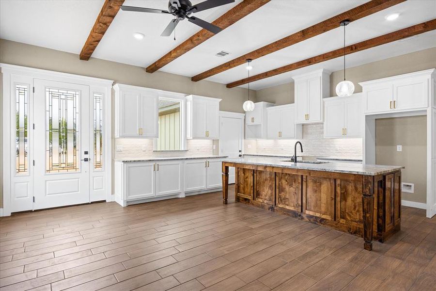 Kitchen featuring white cabinetry, backsplash, a center island with sink, hanging light fixtures, and light stone counters Kitchen featuring white cabinetry, backsplash, a center island with sink, hanging light fixtures, and light stone counters