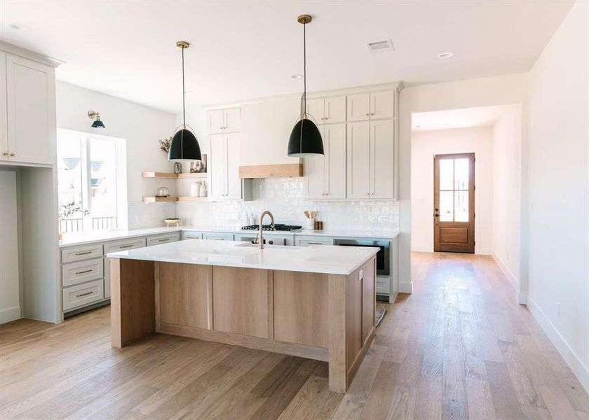 Kitchen with light hardwood / wood-style floors, an island with sink, stainless steel gas stovetop, decorative backsplash, and decorative light fixtures