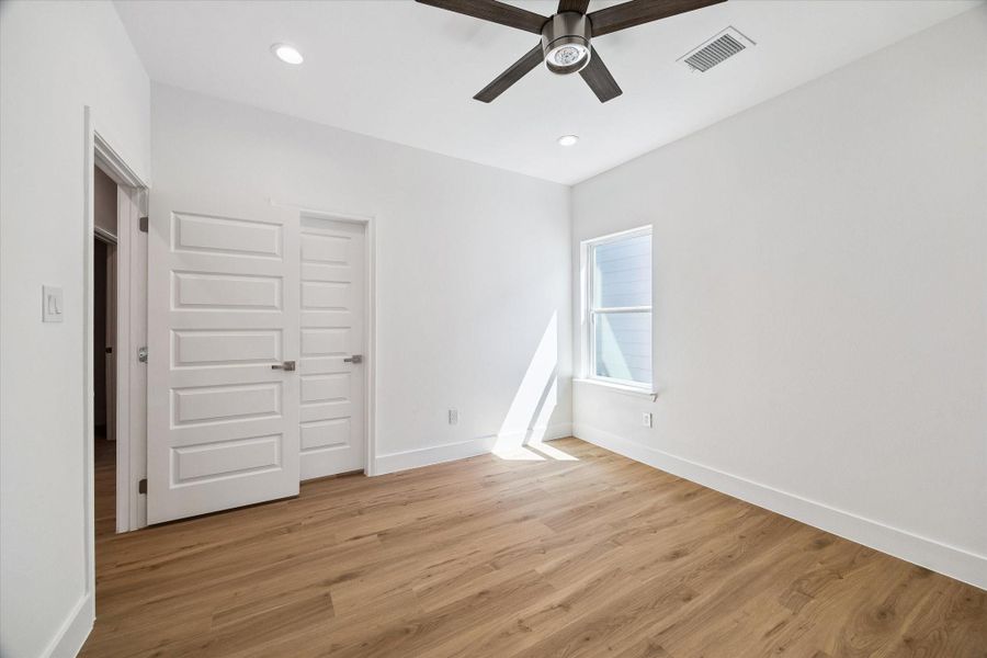 Another view of the secondary bedroom highlights its efficient layout, warm flooring, and crisp modern finishes. Natural light enters through the window, while a contemporary ceiling fan and recessed lighting provide comfort and convenience. Multiple doors offer access to surrounding spaces and storage, while the neutral palette creates an easy backdrop for any decorating style.