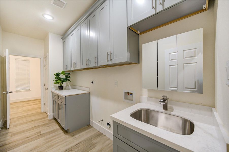 This photo showcases a modern laundry room with sleek gray cabinets, ample countertop space, and a stainless steel sink. The room features light wood flooring and recessed lighting, creating a bright and functional space.