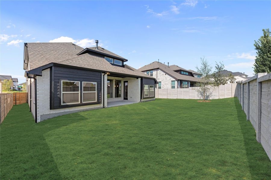 Rear view of two-story home with dark siding, brick exterior, covered patio, and large fenced backyard with green lawn.