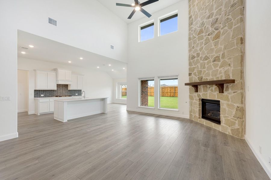 Open living area with two-story ceiling, stone fireplace, hardwood floors, and open kitchen with white cabinets in background Open living area with two-story ceiling, stone fireplace, hardwood floors, and open kitchen with white cabinets in background