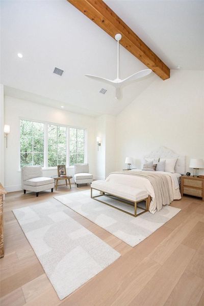 Bright and spacious bedroom with large windows, a vaulted ceiling with wooden beams, and a modern ceiling fan. The room features cozy seating, neutral decor, and light wood flooring, creating a serene and inviting atmosphere.
