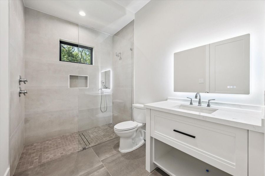 A modern vanity with quartz countertop and undermount sink is paired with a lighted mirror. The walk-in shower, complete with a built-in soap niche, adds both function and style.