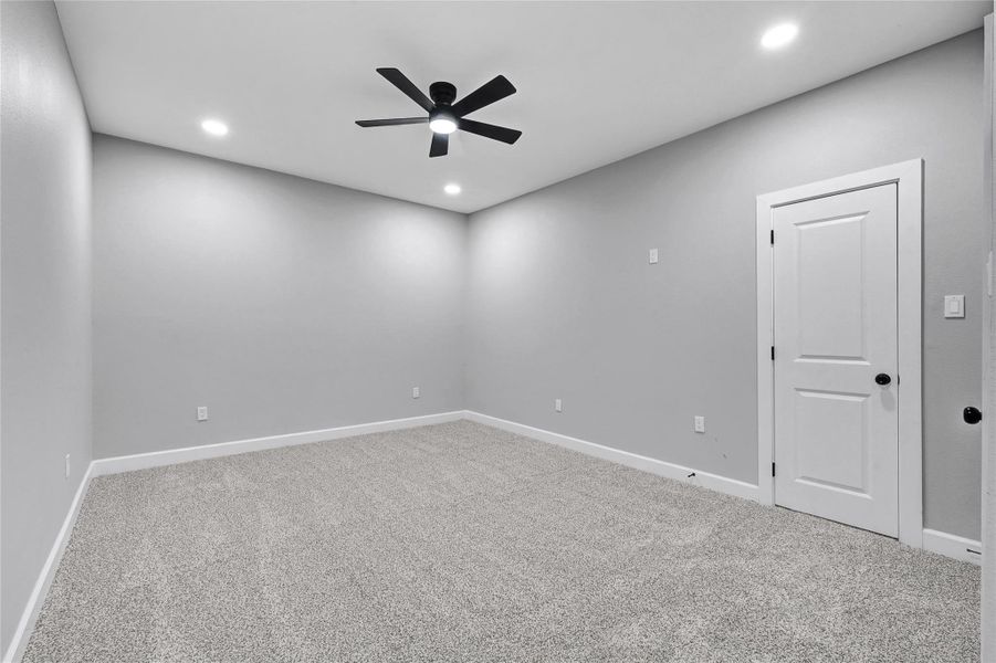 This room features a neutral gray color scheme with plush carpeting, recessed lighting, and a black ceiling fan. It has a simple, modern design with a white door and baseboards, offering a clean and versatile space.