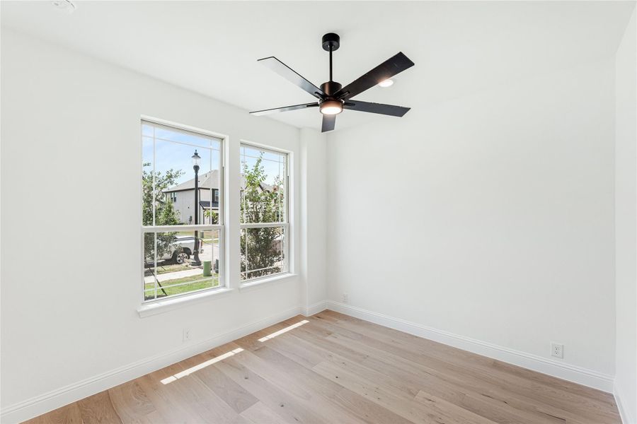 Empty room with light hardwood floors, white walls, black ceiling fan, and two windows overlooking a neighborhood street.