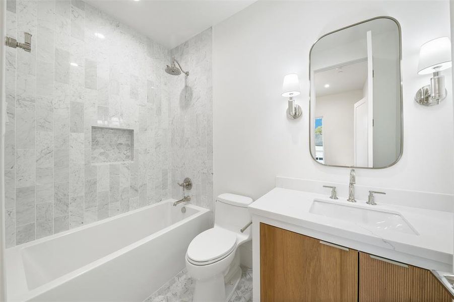 This is a modern bathroom featuring marble wall tiles, a built-in bathtub with a glass shower partition, a sleek vanity with under-sink storage, and stylish wall-mounted light fixtures flanking a rectangular mirror.