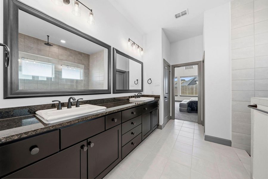 Elegant primary bath showcasing an extended dual vanity with granite countertops, framed mirrors, and modern lighting, complemented by a spacious walk-in shower with sleek tile surround and seamless access to the private suite.