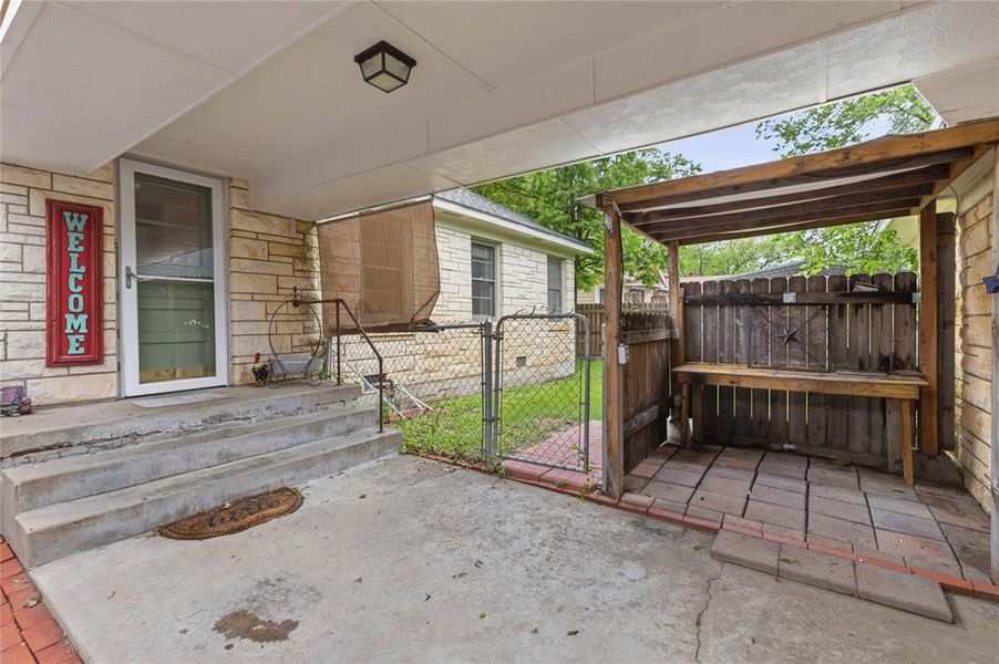 Covered concrete patio featuring stone veneer siding on the exterior, a wooden pergola with a built-in workbench, a metal screen door, and a chain-link fence Covered concrete patio featuring stone veneer siding on the exterior, a wooden pergola with a built-in workbench, a metal screen door, and a chain-link fence