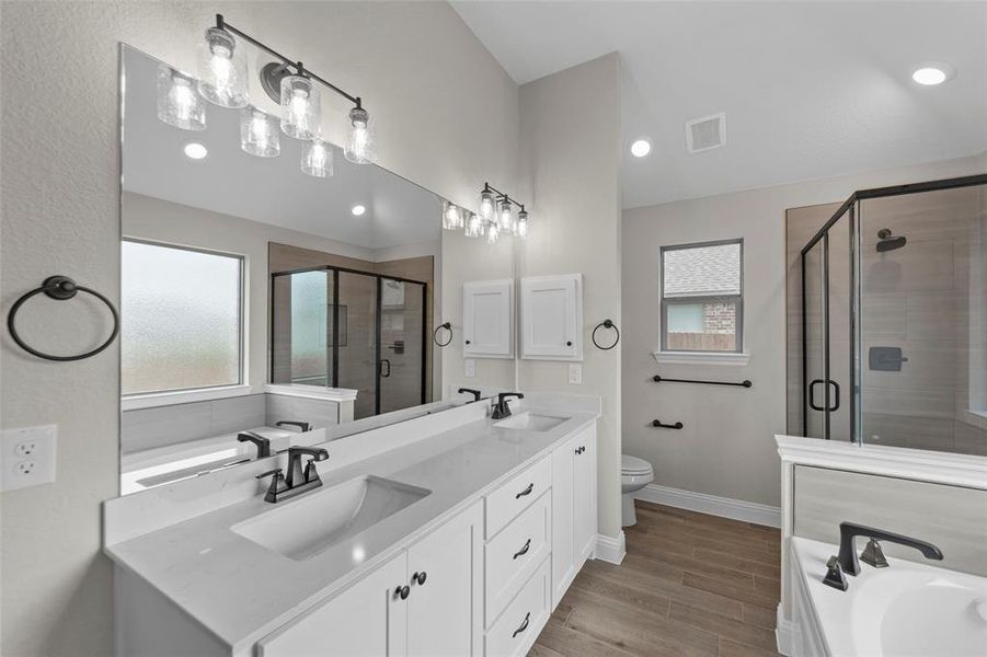 Spacious bathroom featuring a double vanity with stone countertops, a large frameless mirror, a glass-enclosed shower with tile surround, a built-in soaking tub, and wood-finish flooring