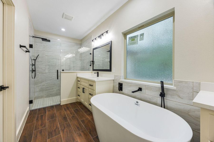 The primary bathroom features a spacious glass-enclosed shower, a sleek freestanding bathtub, and a large frosted window for natural light. The vanity offers ample storage with contemporary fixtures, and the wood-look tile flooring adds a warm touch.