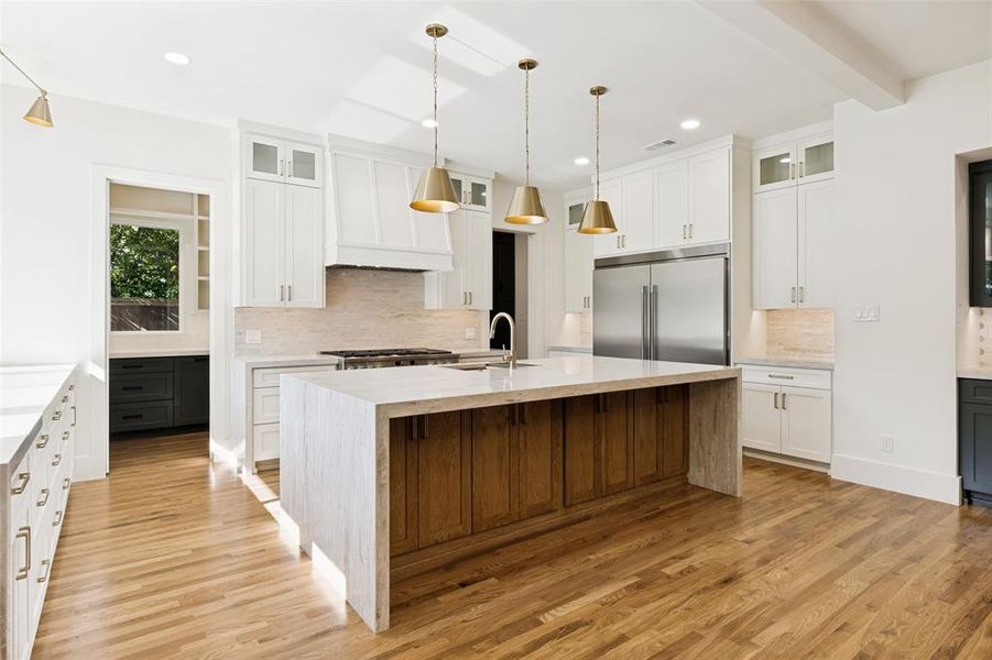 Spacious kitchen featuring a large waterfall island with wood-finish base, stainless steel appliances, white cabinetry, brass pendant lighting, and wood-finish flooring Spacious kitchen featuring a large waterfall island with wood-finish base, stainless steel appliances, white cabinetry, brass pendant lighting, and wood-finish flooring