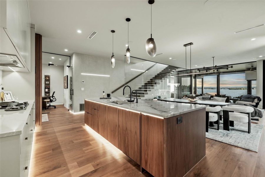 Open-concept living space featuring wood-finish flooring, a kitchen island with integrated lighting, and a glass-paneled staircase