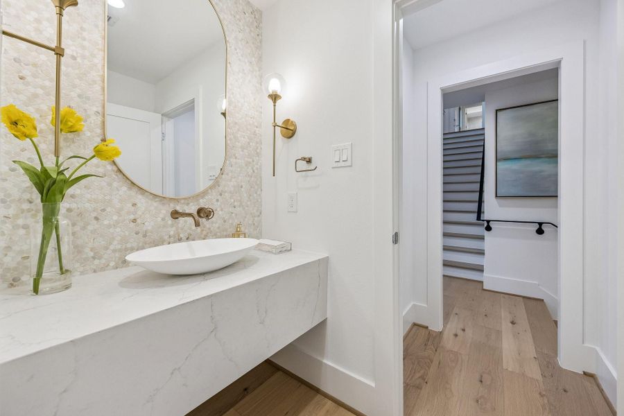 Second Floor Powder Room with a Floating Slab Quartz Counter, Oval Stone Composite White Vessel Sink, Wall-Mounted Widespread Faucet in Champange Bronze. The backsplash wall is adorned with Althena Gold Penny Round Honed Tile. There is also a Oval Framed Mirror and Two Bubble Glass Globe Wall Mounted Sconce Lights in Brass Gold. Second Floor Powder Room with a Floating Slab Quartz Counter, Oval Stone Composite White Vessel Sink, Wall-Mounted Widespread Faucet in Champange Bronze. The backsplash wall is adorned with Althena Gold Penny Round Honed Tile. There is also a Oval Framed Mirror and Two Bubble Glass Globe Wall Mounted Sconce Lights in Brass Gold.