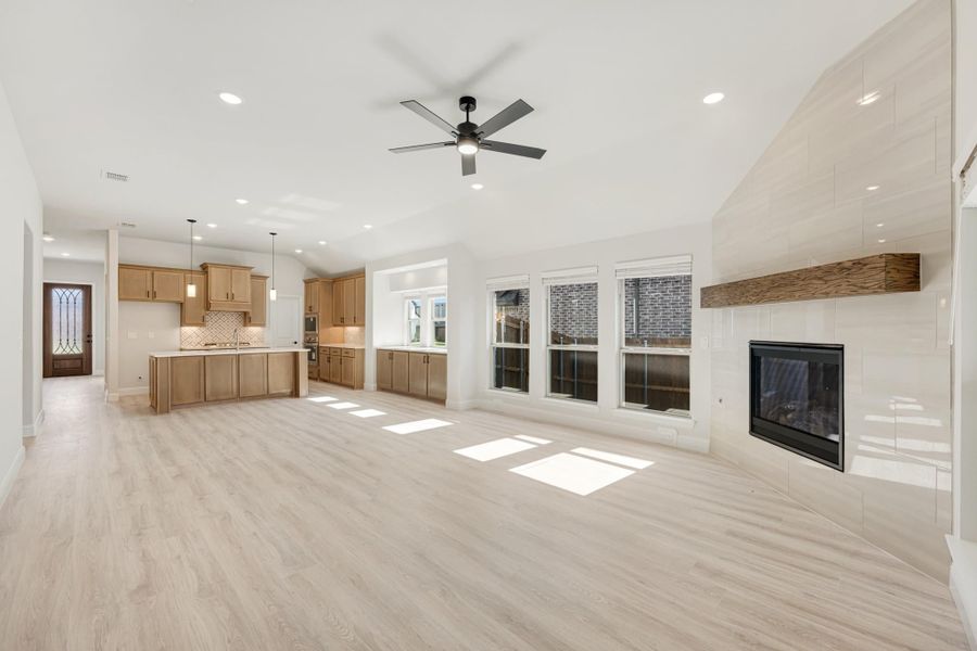 Open living area with light wood floors, tile fireplace, ceiling fan, and kitchen with wood cabinets in background