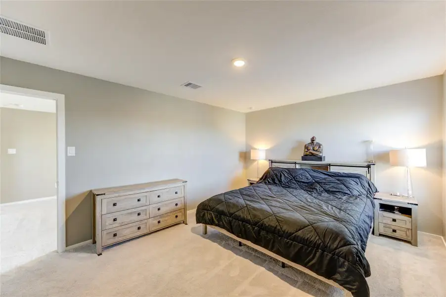 This is a spacious, well-lit bedroom featuring neutral walls, a cozy carpet, and ample space for furniture. It includes a large bed, a chest of drawers, and bedside tables with modern lamps, giving it a comfortable and inviting feel.